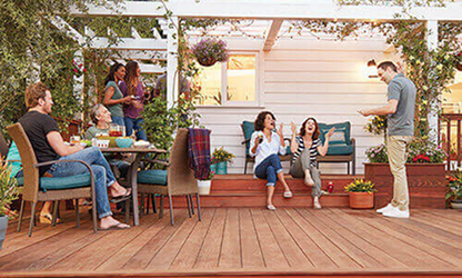 Group of six friends socializing on a wooden deck with chairs and plants under a pergola at sunset.