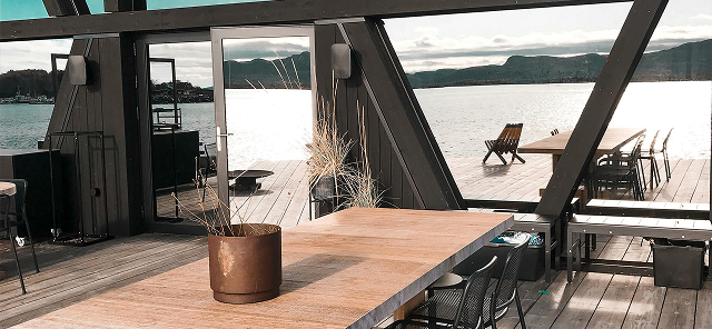 Modern indoor seating area with wooden table, black chairs, and glass walls overlooking a lake and mountains.