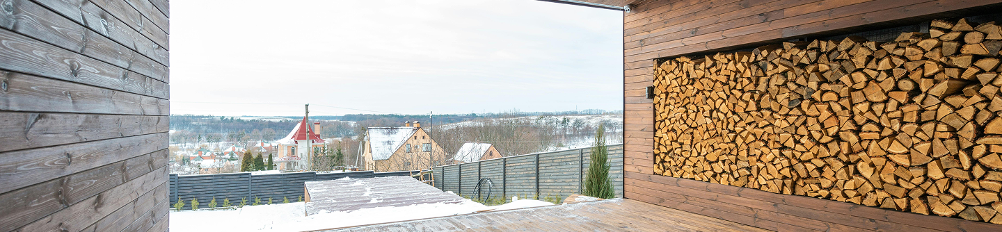 Snow-covered backyard with wooden deck and neatly stacked firewood on a wooden wall panel.