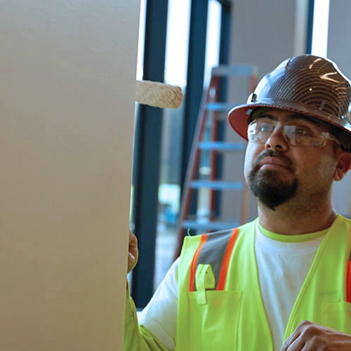 Construction worker wearing a hard hat, safety glasses, and a neon yellow safety vest painting a wall indoors.
