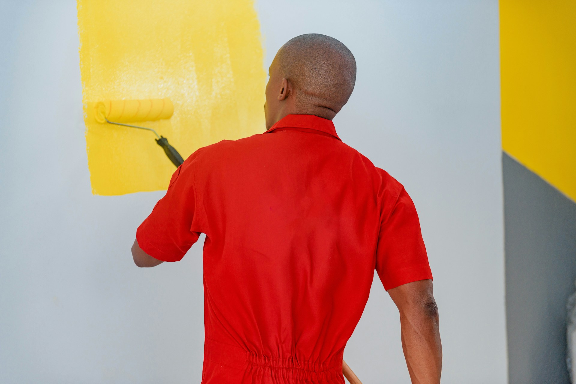 Man in red coveralls painting a wall yellow with a paint roller.