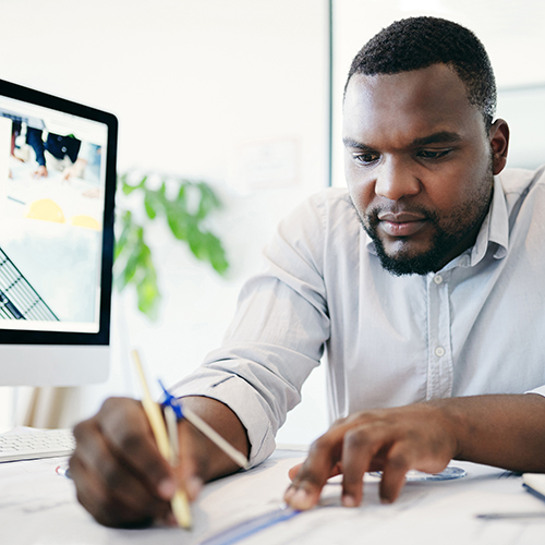 Man in a light shirt focused on drawing with a pencil at a desk with a computer and architectural plans.
