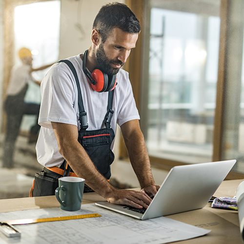 Construction worker wearing headphones around his neck working on a laptop at a site with blueprints on the table.
