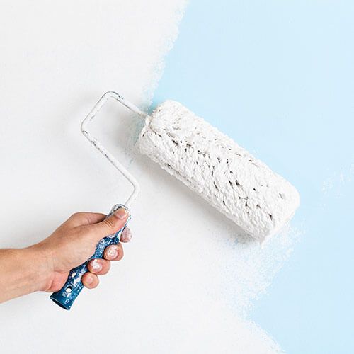 Hand holding a paint roller applying white paint over a blue wall.