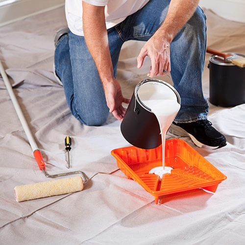 Person kneeling on a tarp pouring white paint from a black bucket into an orange paint tray with a roller and screwdriver nearby.