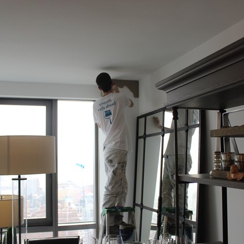 Painter on a ladder applying paint to an upper corner wall in a modern apartment with large windows and city view.
