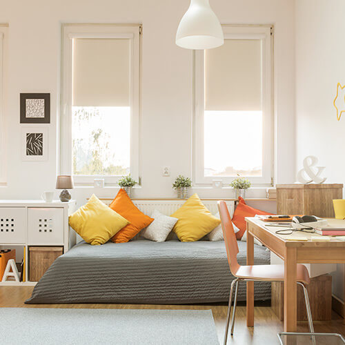 Bright living room with a gray sofa covered by colorful yellow and orange pillows, wooden desk with chair, and two large windows with blinds.