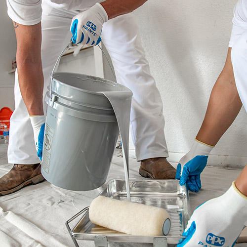 Two painters wearing white clothes and gloves pouring gray paint from a bucket into a paint tray with a roller on a drop cloth.