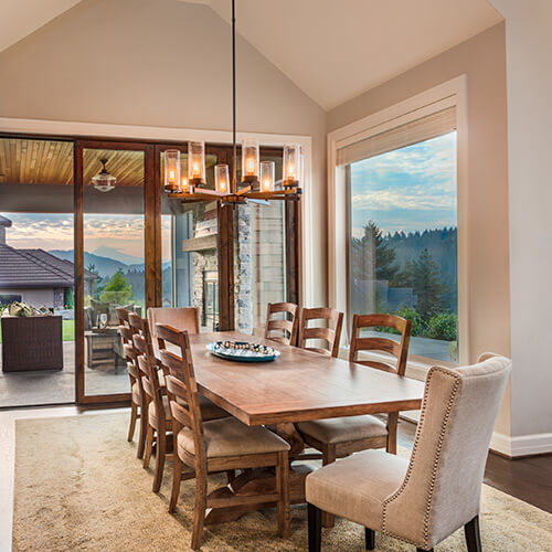 Modern dining room with wooden table, six wooden chairs, one upholstered chair, large windows showing trees and mountains at sunset.