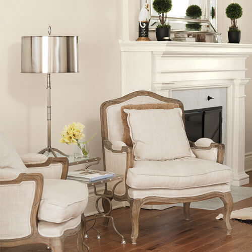 Cozy living room with two beige cushioned armchairs, metal floor lamp, small glass side table with yellow flowers, and white fireplace mantel with decorative plants.