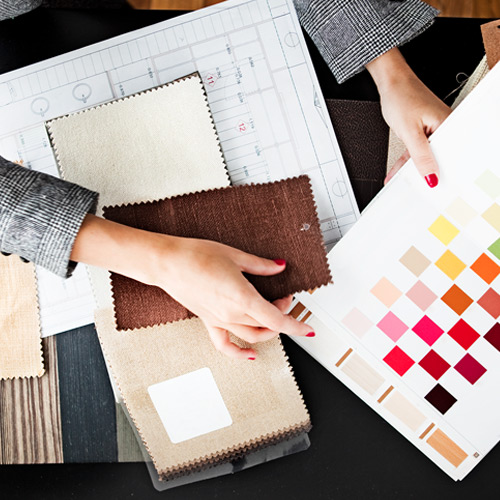 Hands holding fabric swatches and a color palette chart over architectural blueprints on a table.