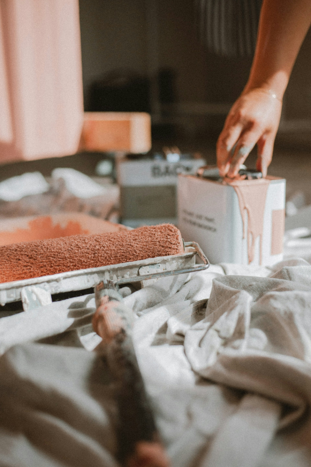 Close-up of a paint roller covered in peach paint lying on a cloth with a hand dipping a brush into a paint can.