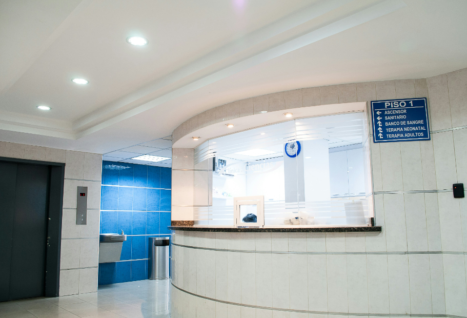Hospital or clinic waiting area with reception window, elevator doors, drinking fountain, blue tiled wall, and directional sign in Spanish.