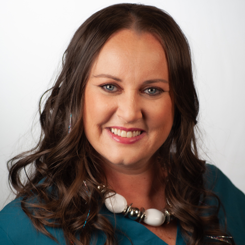 Smiling woman with long curly dark hair wearing a teal top and a chunky white and silver necklace.