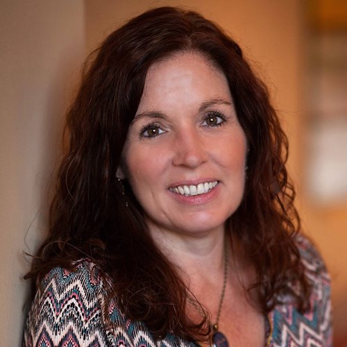 Smiling woman with long curly brown hair wearing a patterned top and a necklace, standing indoors with a blurred background.