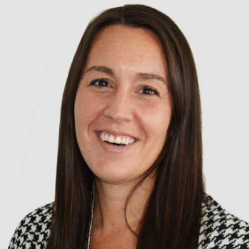 Smiling woman with long brown hair wearing a black and white patterned jacket against a gray background.