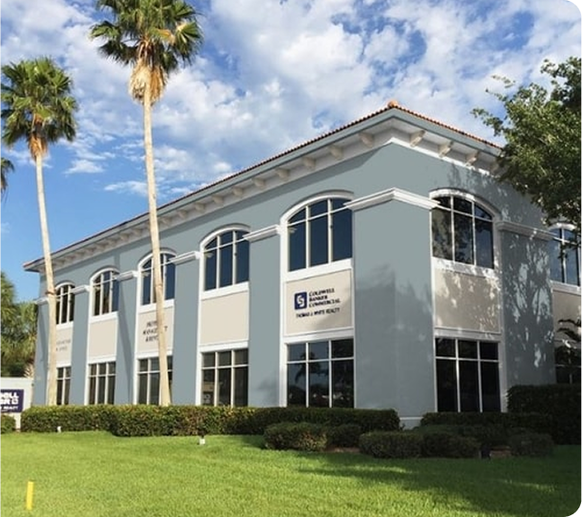 Two-story office building with blue-gray and beige walls, large arched windows, surrounded by green lawn, palm trees, and shrubs under a partly cloudy sky.