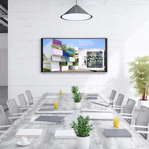 Modern conference room with a long wooden table, white chairs, potted plants, notebooks, orange juice glasses, and a wall-mounted screen displaying architectural visuals.