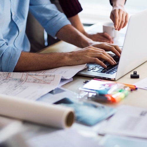Two people working together on a laptop surrounded by architectural plans and stationery.