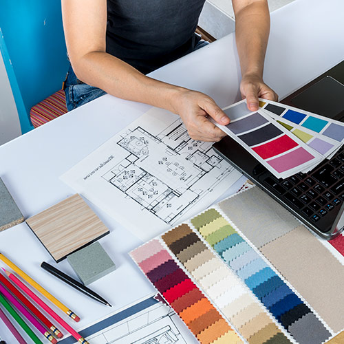 Person reviewing color swatches and architectural floor plans on a desk with pencils, pen, wood samples, and a laptop.
