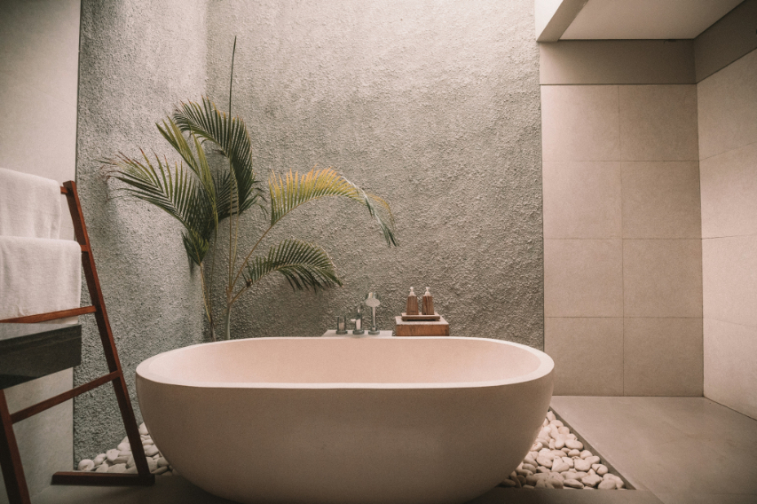 Minimalist bathroom with a freestanding oval bathtub surrounded by white pebbles, a textured gray wall, a wooden towel rack with white towels, and a tall green plant.