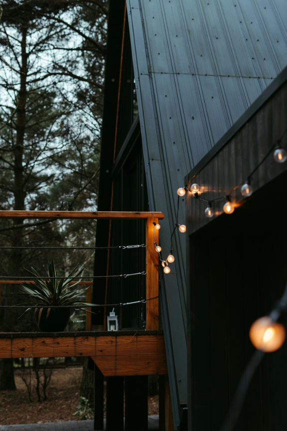 String lights glowing softly along the edge of a wooden deck attached to a dark exterior cabin in a forested area.