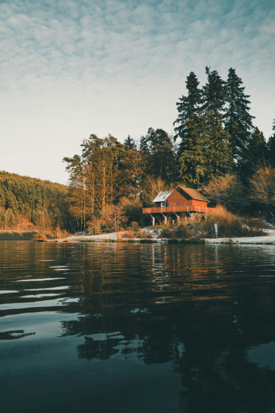 Red cabin on stilts by a calm lake surrounded by tall trees in soft evening light.