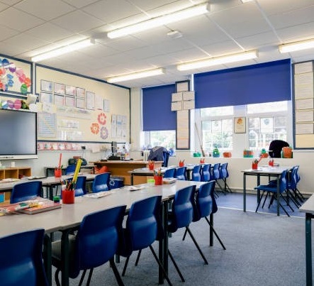Classroom with rows of blue chairs and tables, colorful pencil holders, educational posters on walls, and blue window blinds.