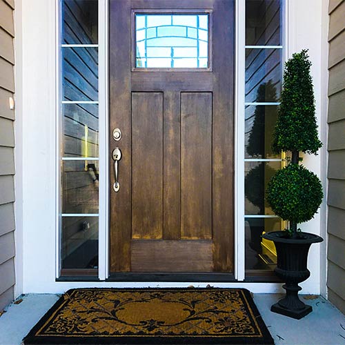 Dark wood front door with a decorative window panel and glass sidelights, flanked by a potted topiary and a brown patterned doormat.