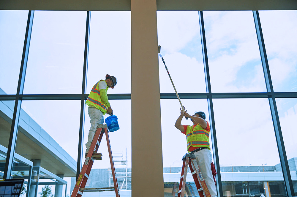 Two workers in safety vests and helmets painting an interior wall near large windows, each standing on separate ladders.