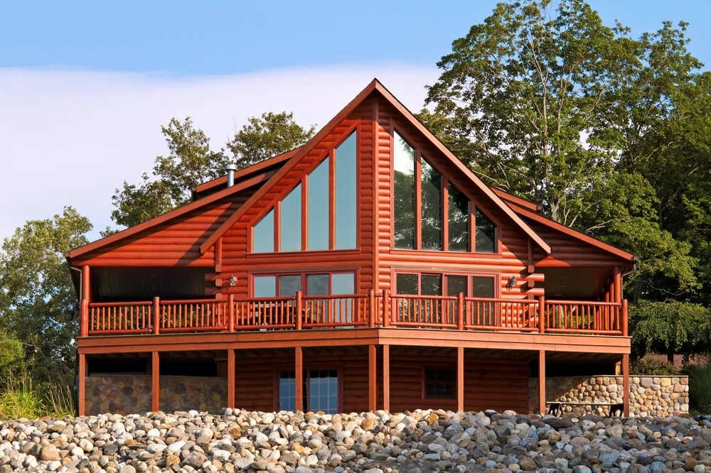 Red wooden cabin with large glass windows and a wraparound porch surrounded by trees and rocks in front.