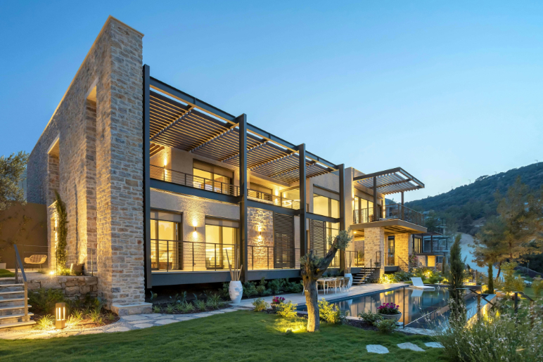 Modern two-story stone and glass house with warm interior lights, a swimming pool, and surrounding greenery at dusk.