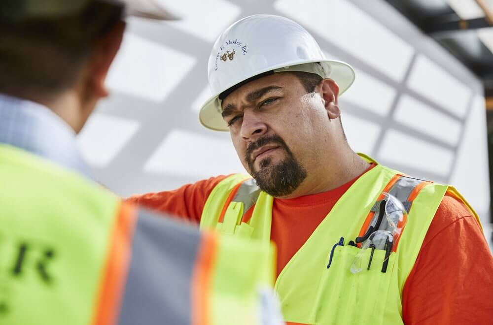 Construction worker wearing a white hard hat and yellow safety vest talking to another worker at a construction site.