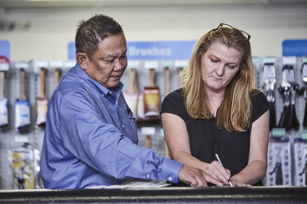 Man in blue shirt assisting woman in black shirt signing a document in a store aisle with brushes in the background.