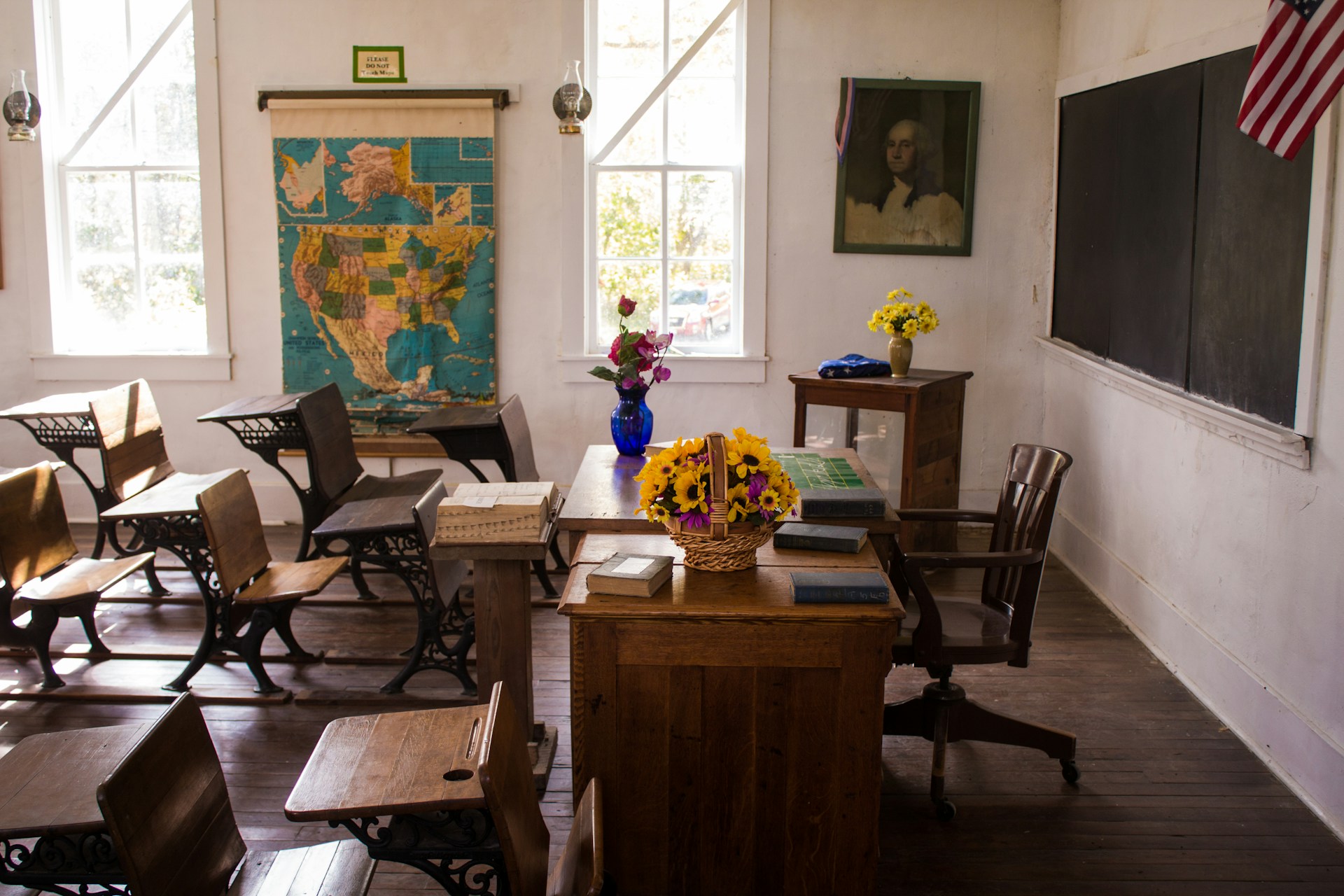 Empty vintage classroom with wooden desks, a teacher's desk adorned with flower baskets, a US map on the wall, and a portrait of George Washington.