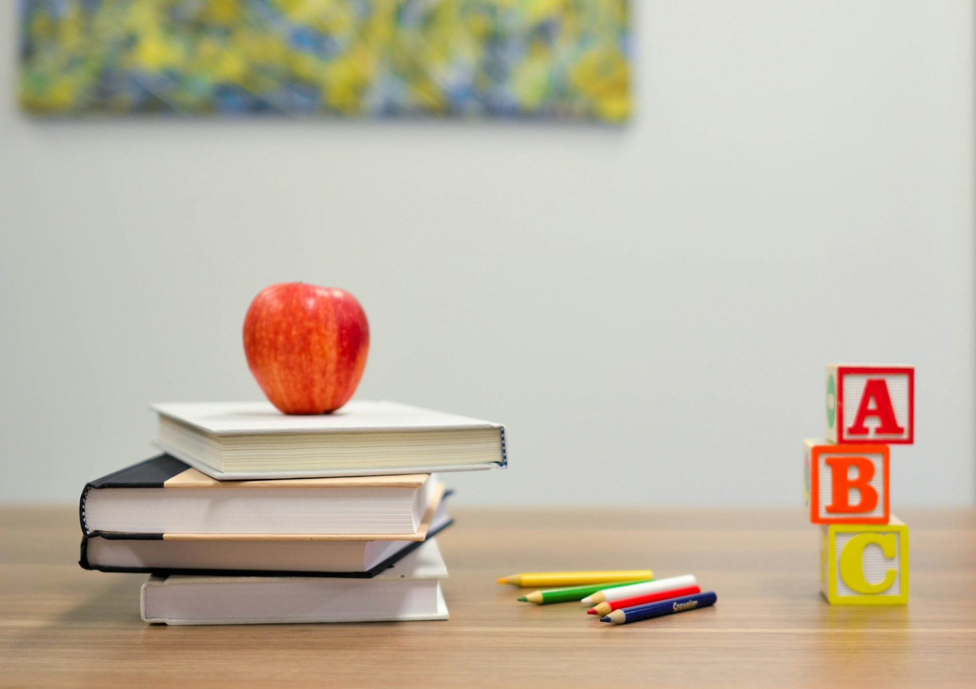 Red apple on top of a stack of books next to colored pencils and ABC alphabet blocks on a wooden table.