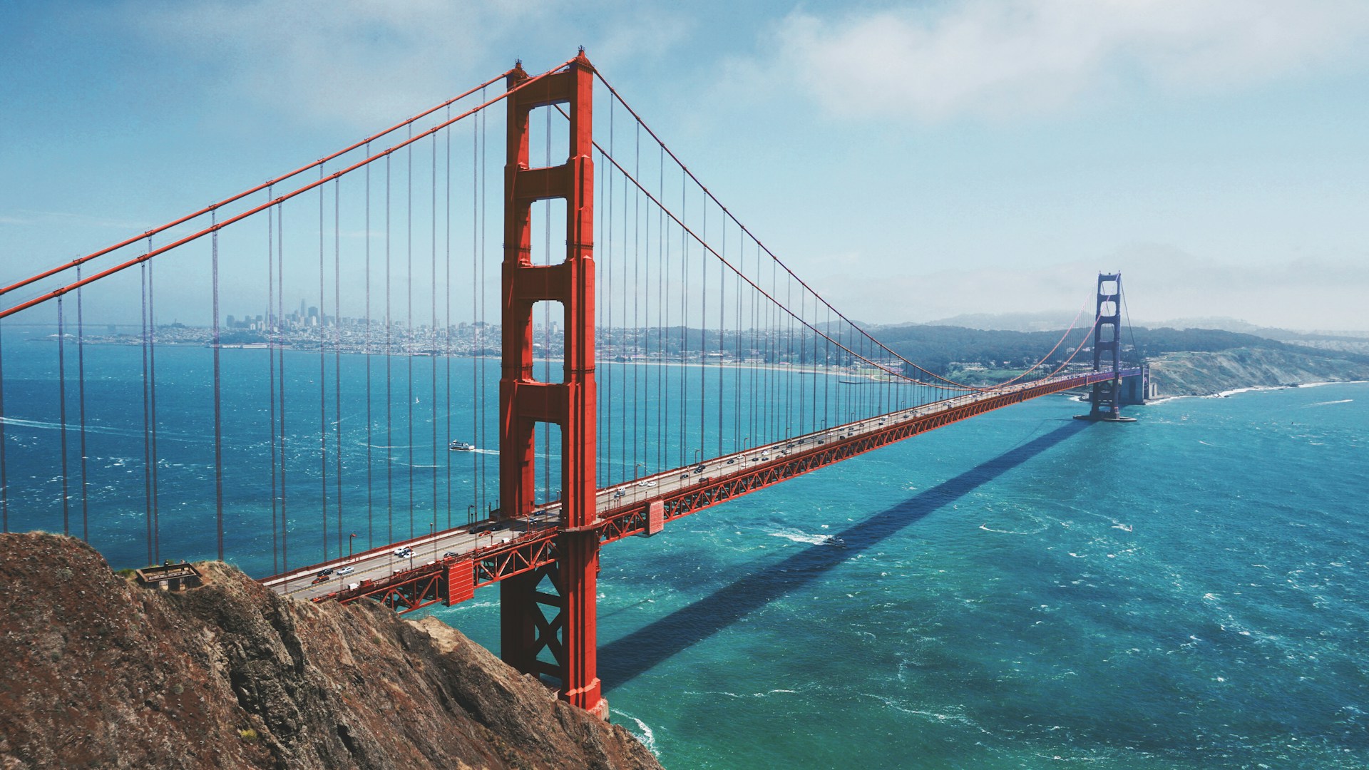 Wide-angle view of the Golden Gate Bridge spanning blue water on a clear day with city and hills in the background.