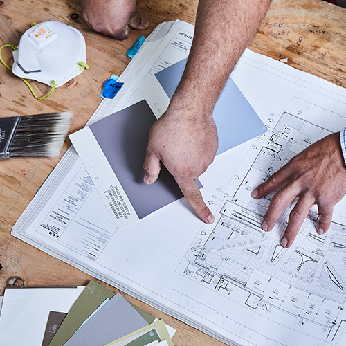 Hands of a person pointing at a blueprint on a wooden table surrounded by paint swatches, a paintbrush, and a protective face mask.