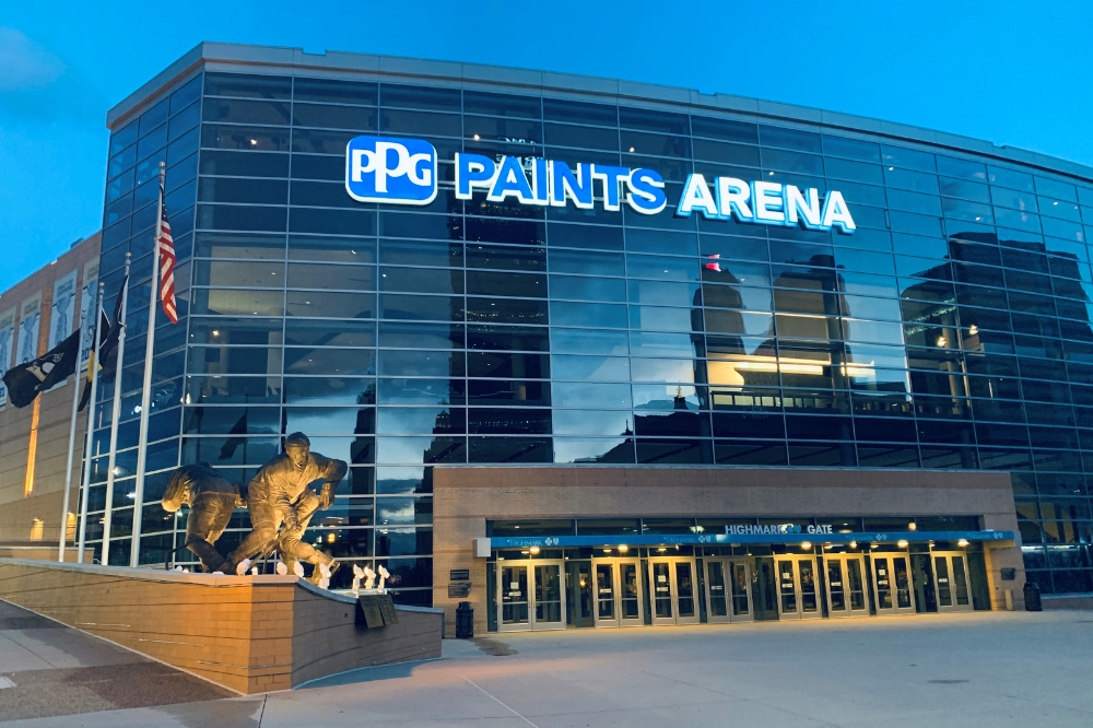 Exterior of PPG Paints Arena at dusk with statues of hockey players and flags on poles.