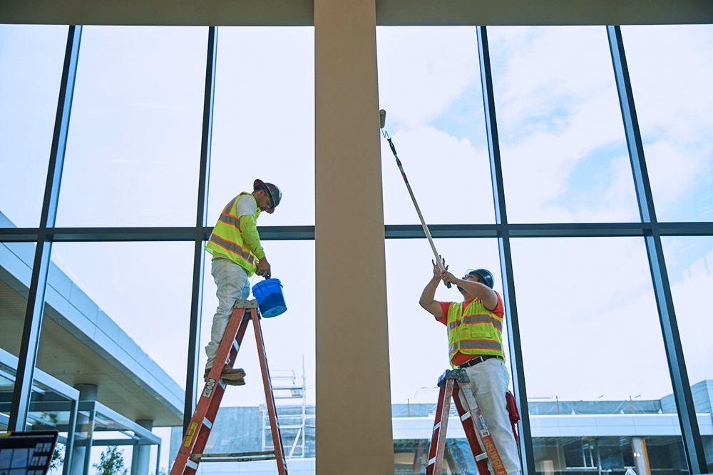 Two painters wearing safety vests and helmets, standing on ladders, painting a tall interior column near large glass windows.
