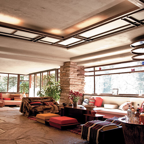 Spacious living room with large windows, stone pillar, modern ceiling lights, and cozy seating including sofas and floor cushions.