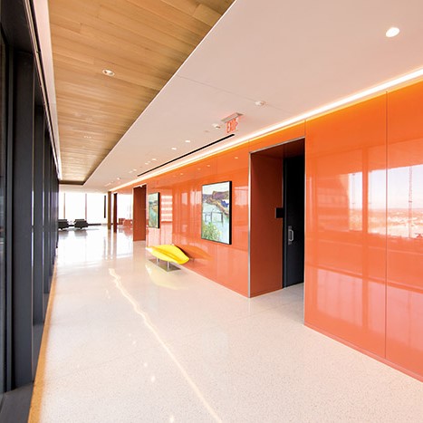 Modern office hallway with glossy orange wall panels, a yellow bench, large windows, and polished white flooring.