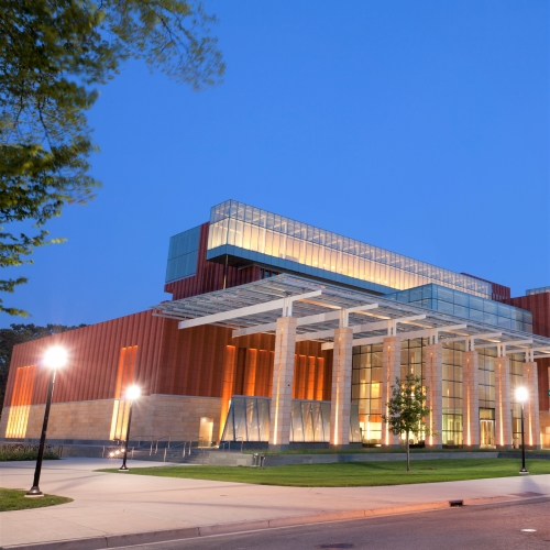 Modern building with glass and stone facade illuminated at dusk with street lamps and a tree nearby.