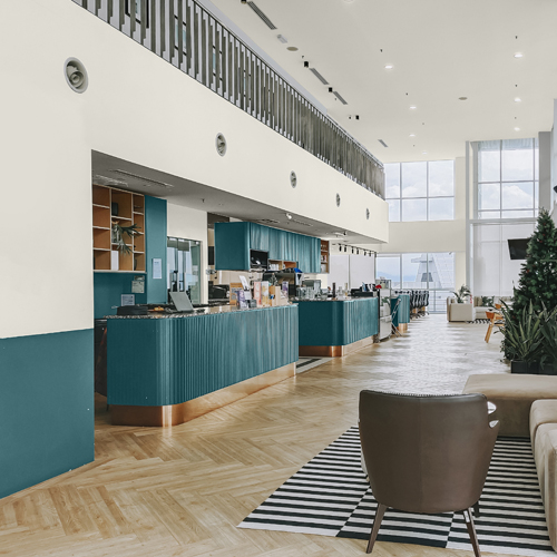 Modern café interior with teal counters, light wood flooring, large windows, and seating area including a brown chair on a striped rug.