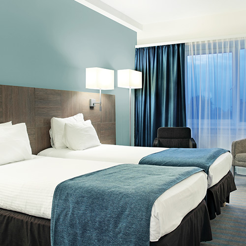 Hotel room with two twin beds featuring white linens, blue throws, wooden headboard, wall-mounted lamps, and blue curtains by a window.