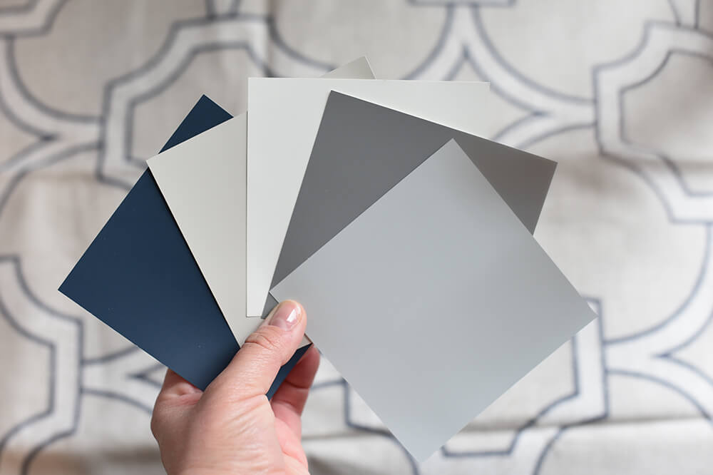 Hand holding five paint color sample cards in shades of blue, gray, and beige against a patterned fabric background.