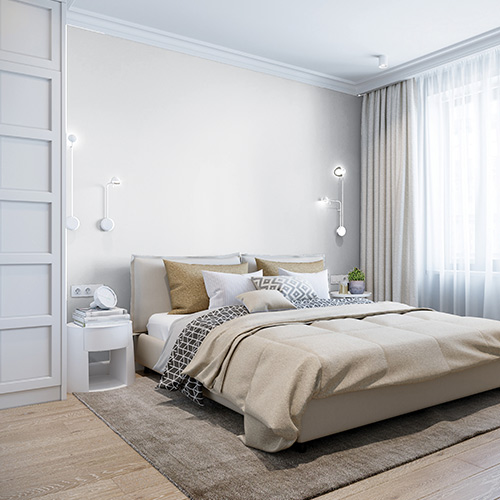 Modern bedroom with beige bedding, pillows, wall-mounted reading lights, a nightstand with books and clock, and sheer curtains letting in natural light.
