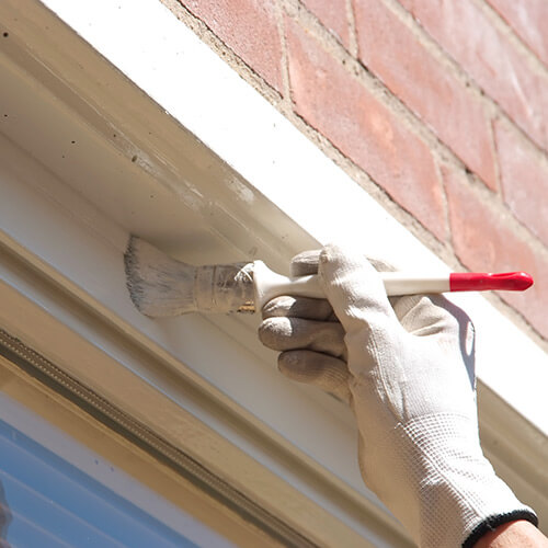 Hand wearing white glove painting white trim on building exterior with a paintbrush.