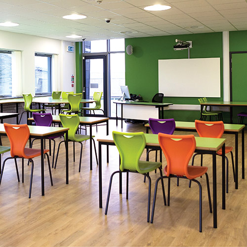 Modern classroom with colorful orange, green, and purple chairs and wooden tables arranged neatly, featuring a green accent wall with a whiteboard and projector.