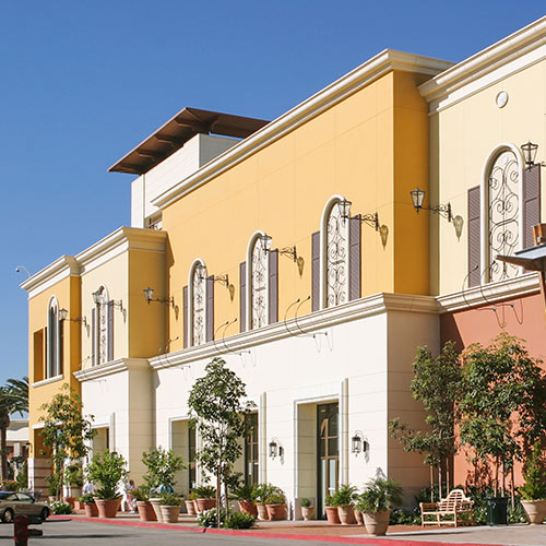 Exterior of a colorful retail building with large arched windows, decorative ironwork, potted plants, and clear blue sky.